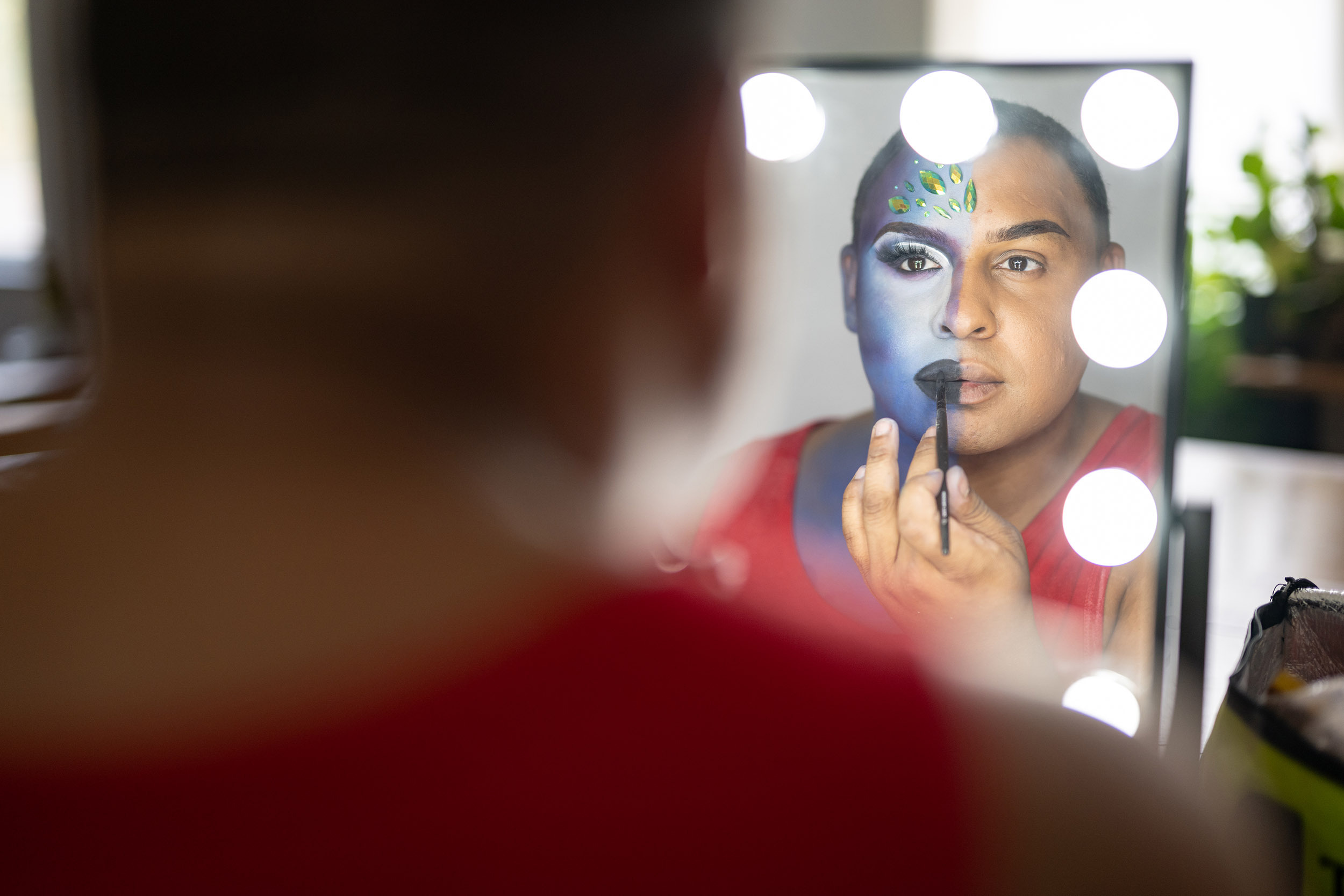 Reflection of Voyages Featured Artist Devon Vaow Applying Blue Makeup to One Side of His Face in a Mirror